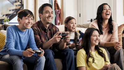A family of five sits on the couch, smiling and playing together. Four of the family members are holding controllers.